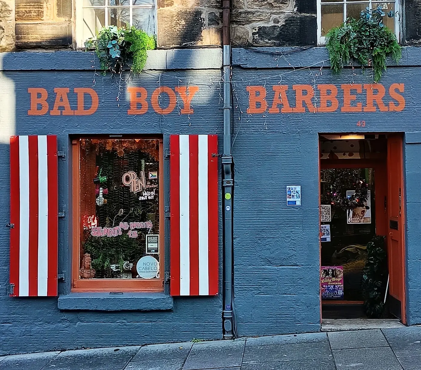 Bad Boy Barbers shopfront on Candlemaker Row, Edinburgh — orange fascia lettering, red and white striped shutters and ferns over the window